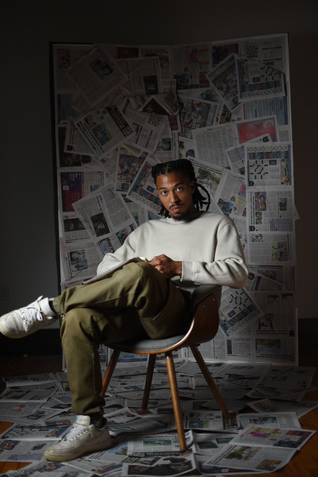 Artistic portrait on chair surrounded by newspaper collage wall in dim lighting.