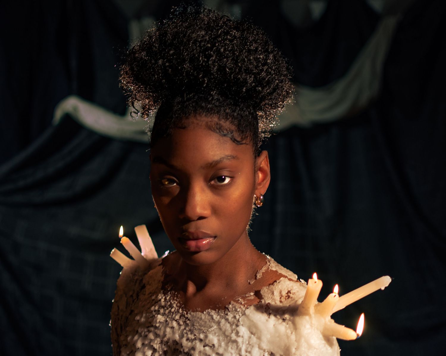 Young Black woman with natural updo hair, wearing ornate costume with candles, posed against dark dramatic backdrop.