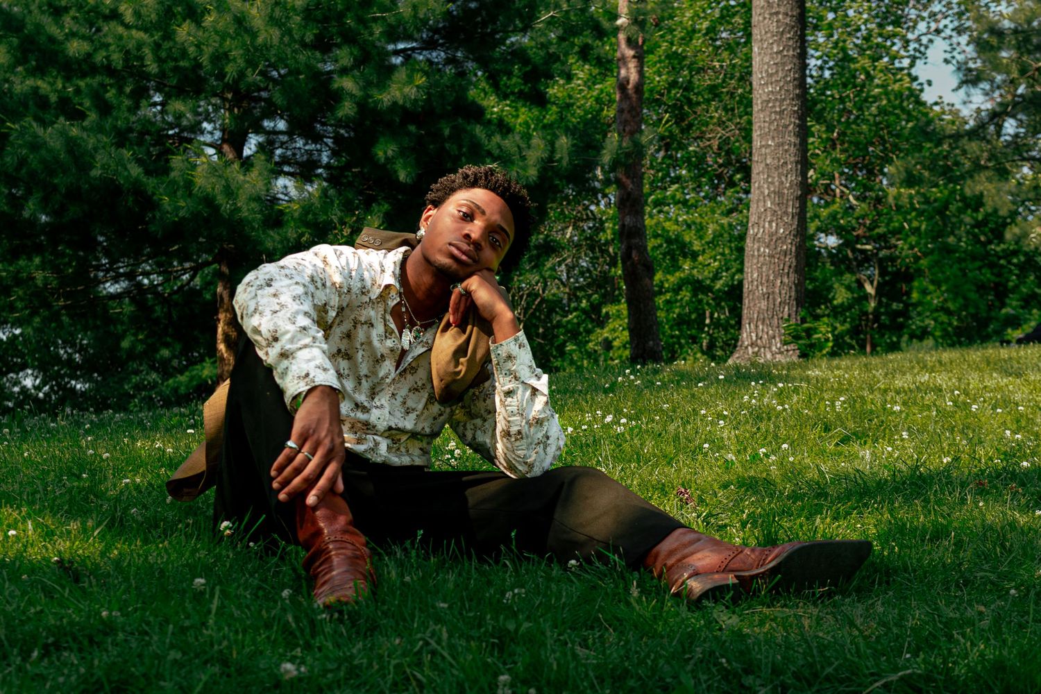 Person in floral shirt and brown boots sitting on green grass in a park, gazing upward thoughtfully.