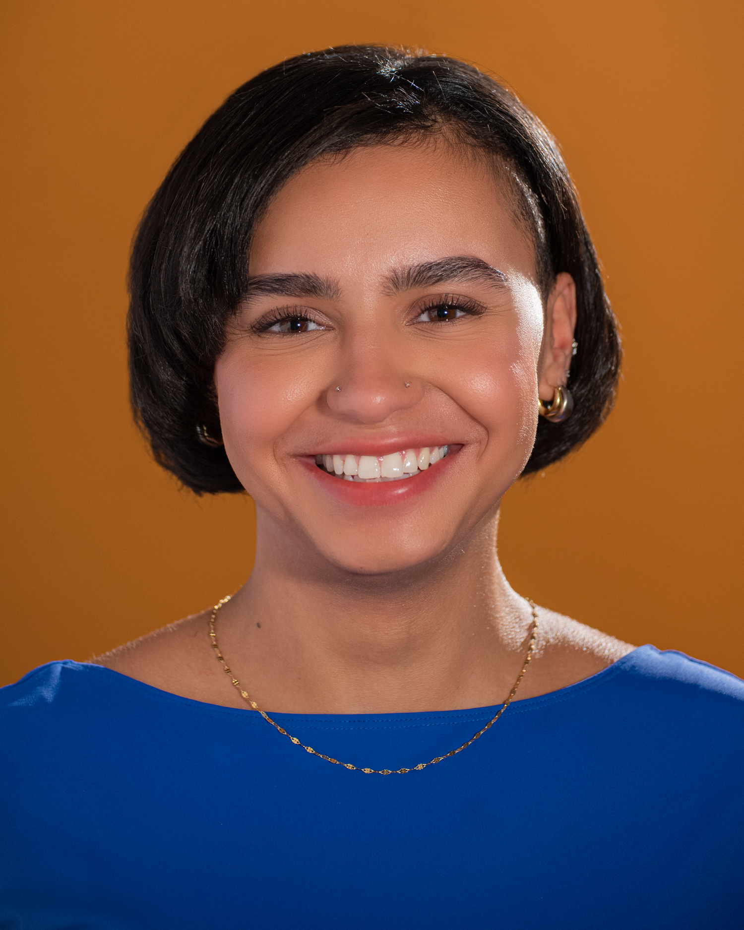 Bright portrait against orange background wearing royal blue top with delicate jewelry.
