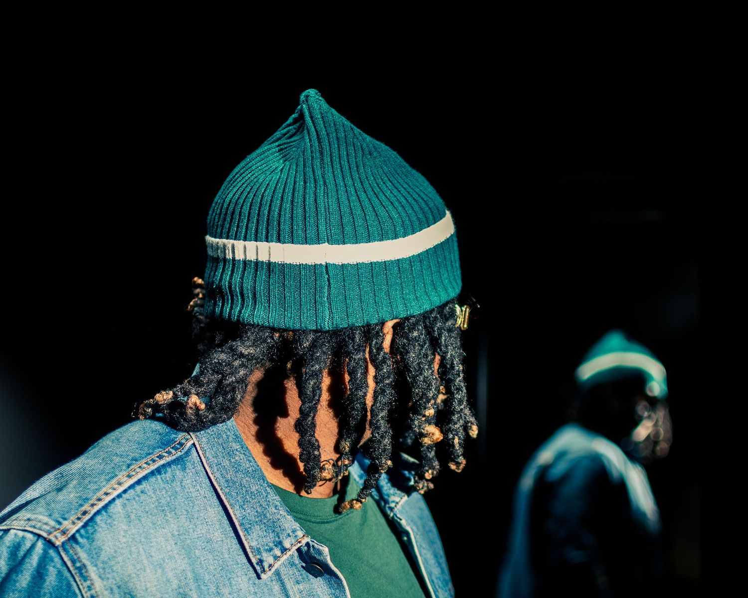 Person in teal knit beanie and denim jacket shown in profile against black backdrop.