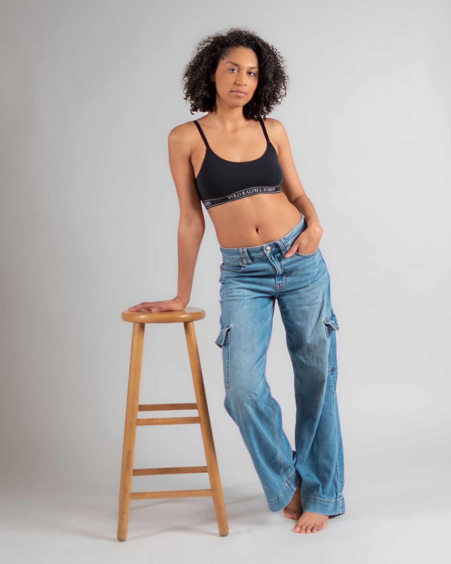 Woman wearing black sports bra and light wash cargo jeans, posing next to a wooden stool against a gray background.