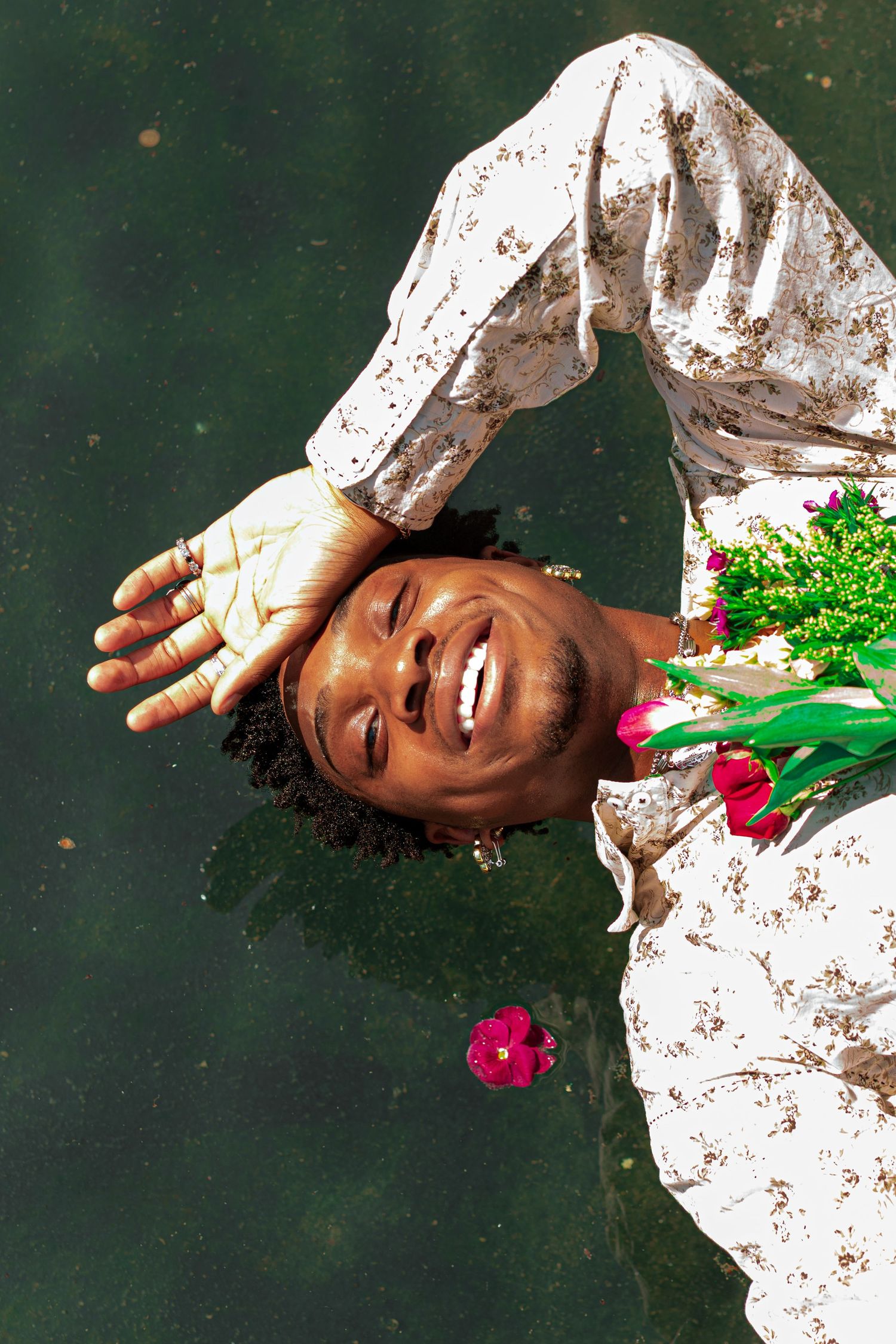 Smiling Black man floating in water wearing a floral shirt, holding pink roses and green flowers, hand raised near face.
