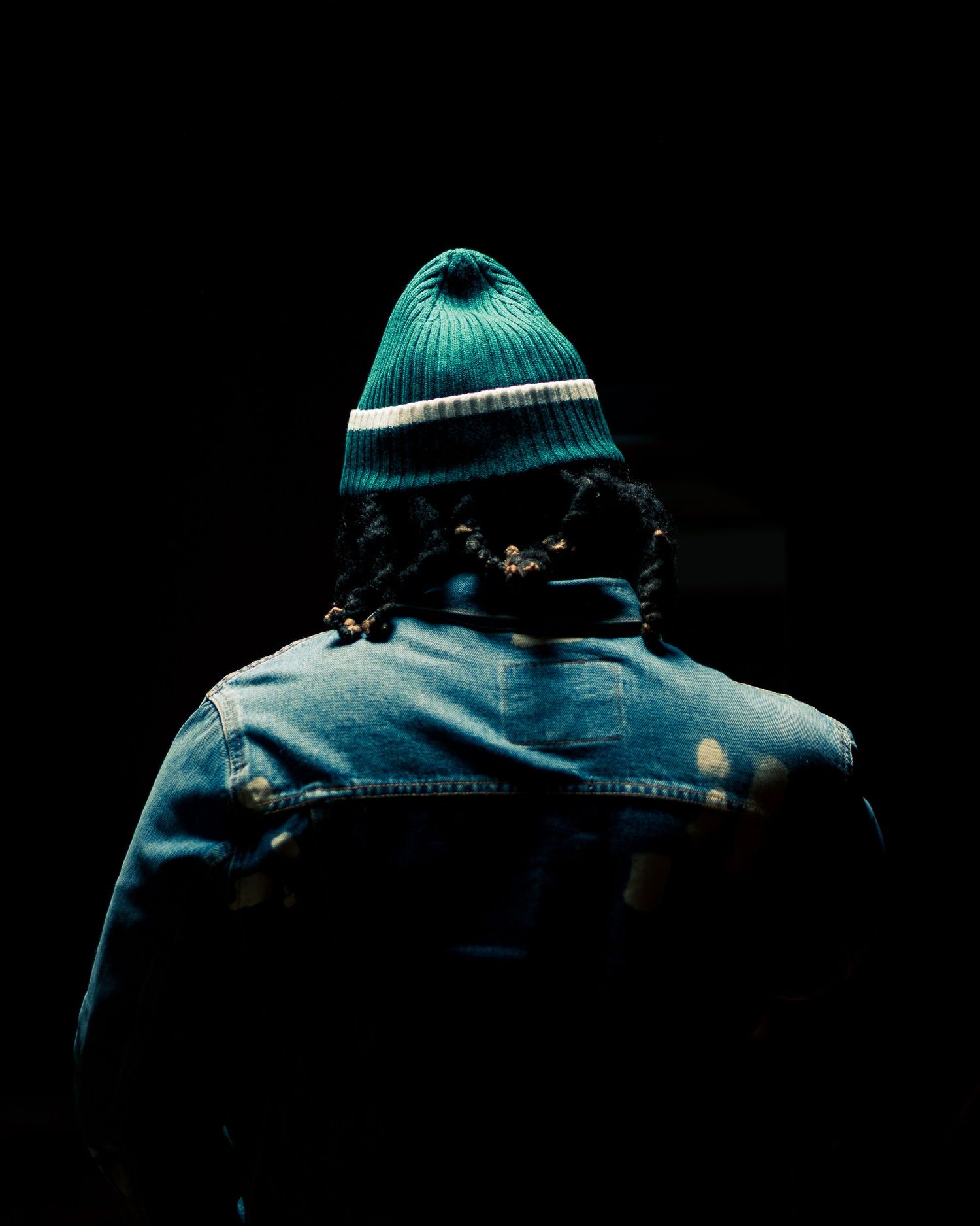 Silhouette of person wearing teal beanie and denim jacket against dark background.