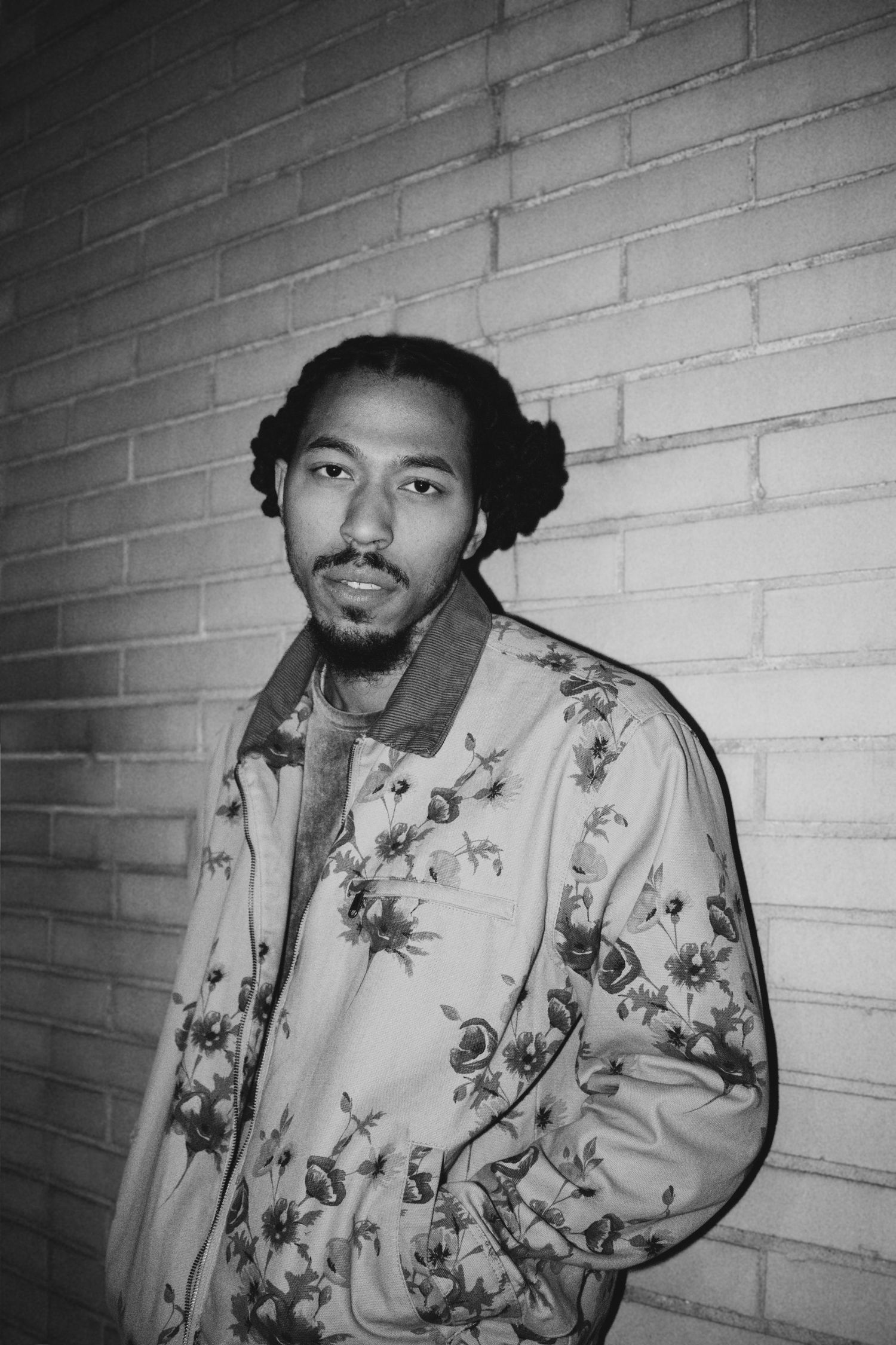 Black and white portrait of someone wearing a floral patterned jacket against a brick wall.