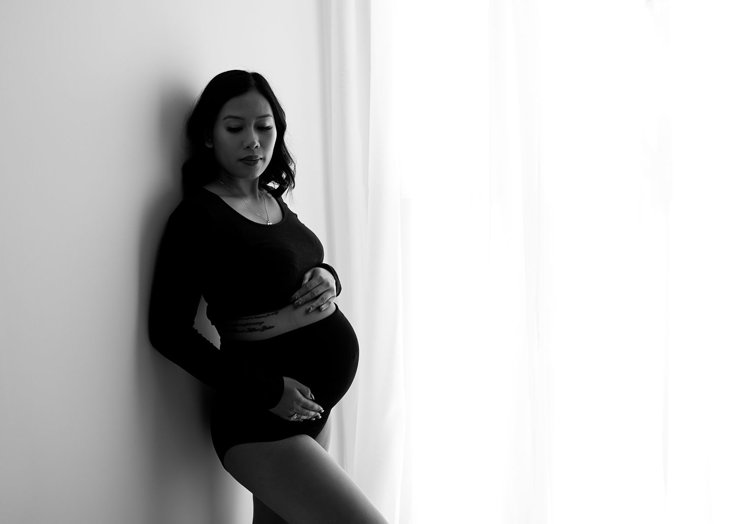 Black and white pregnancy photo. Subject holding belly while leaning on studio wall.