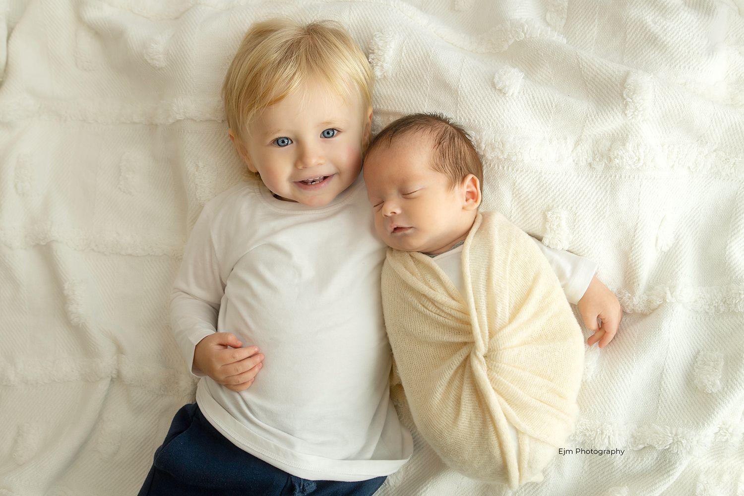 Baby boy laying on a nice blanket holding his new newborn sibling.