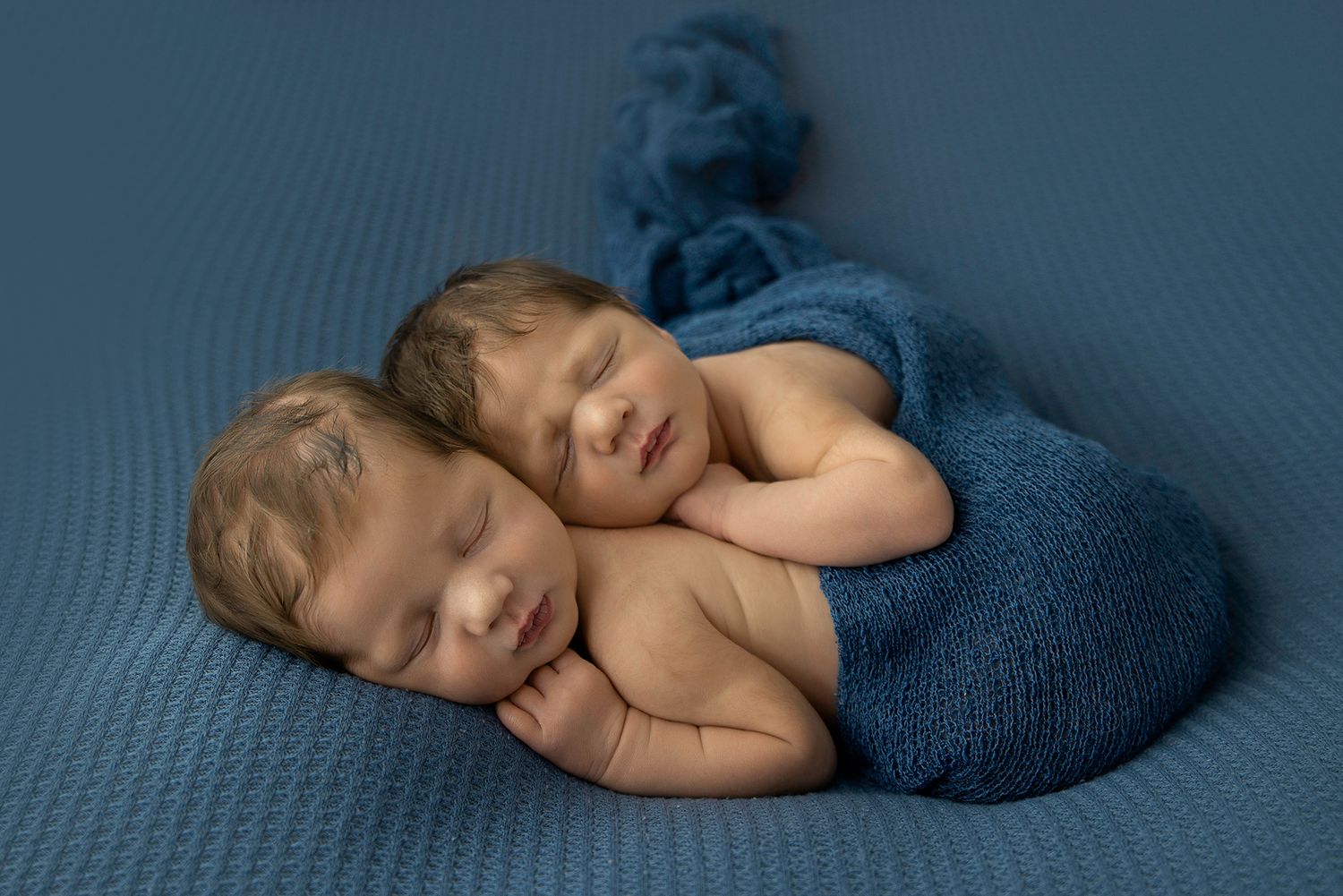 Newborn twin brothers posed together on a blue blanket and wrap sleeping closely.