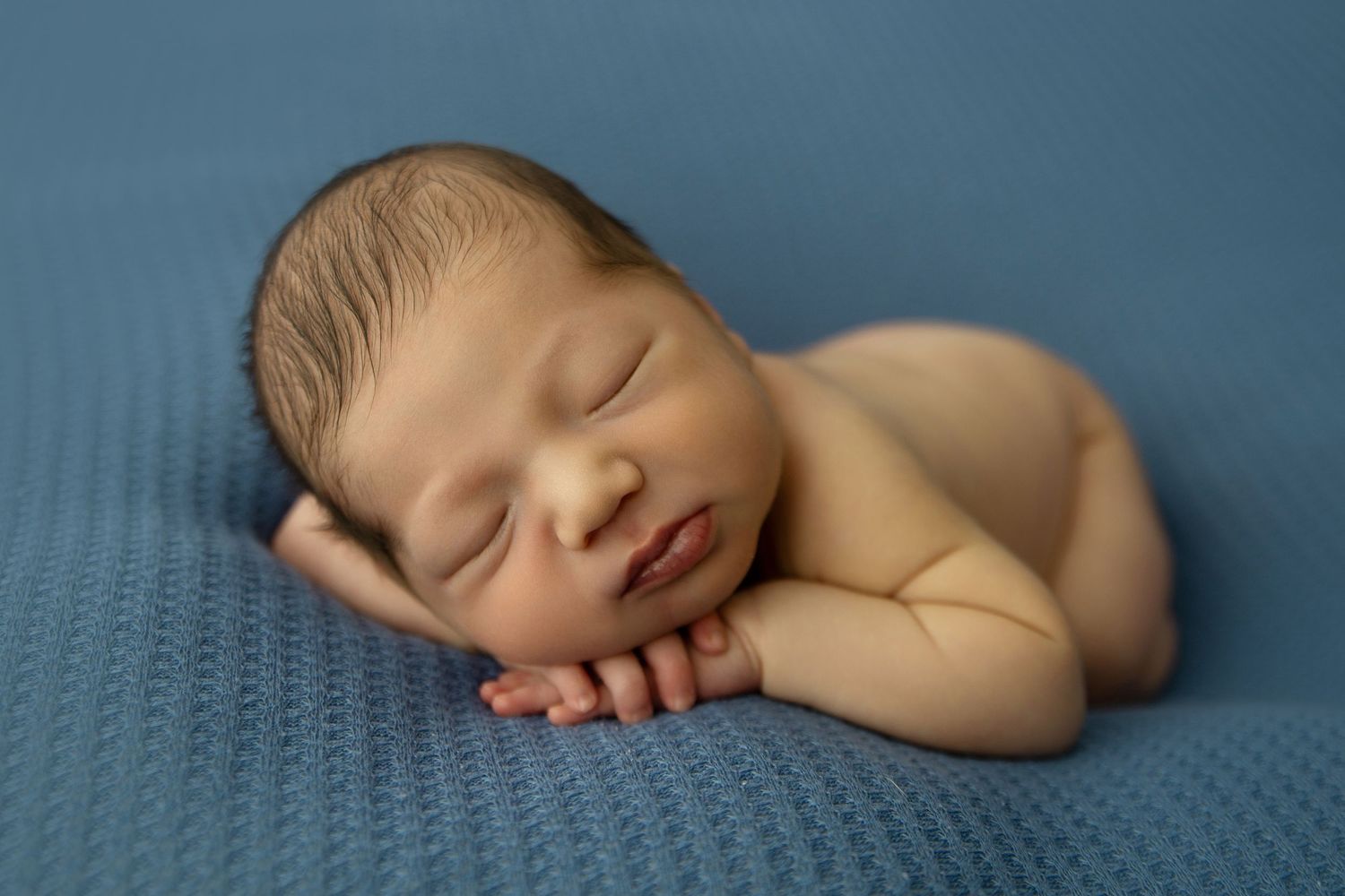 Newborn baby boy posed with chin resting on both hands on a navy blue blanket.
