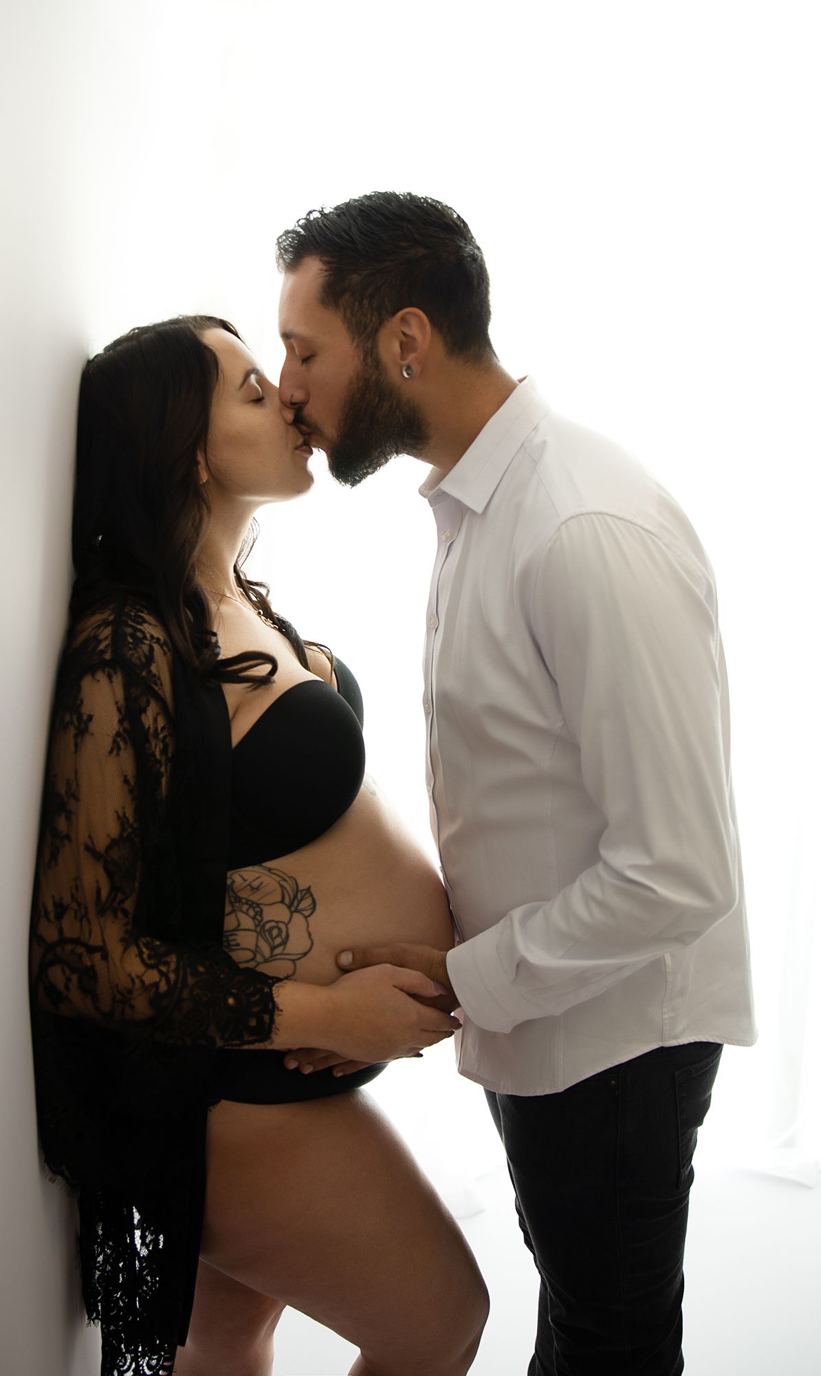 Pregnant woman leaning against the awl in a rode with pregnant belly exposed, husband leaning in towards her, holding her belly kissing her.