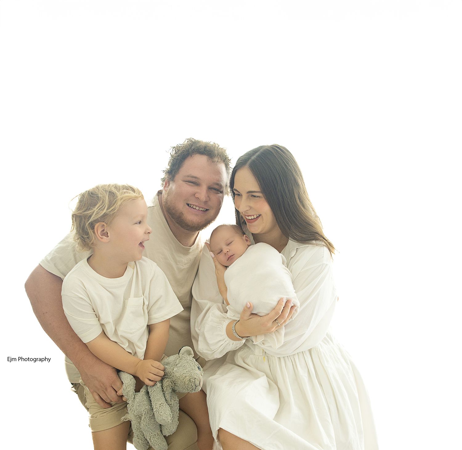 Family photo all in white of parents with newborn baby and their older son. Older son holding tightly to his teddy.
