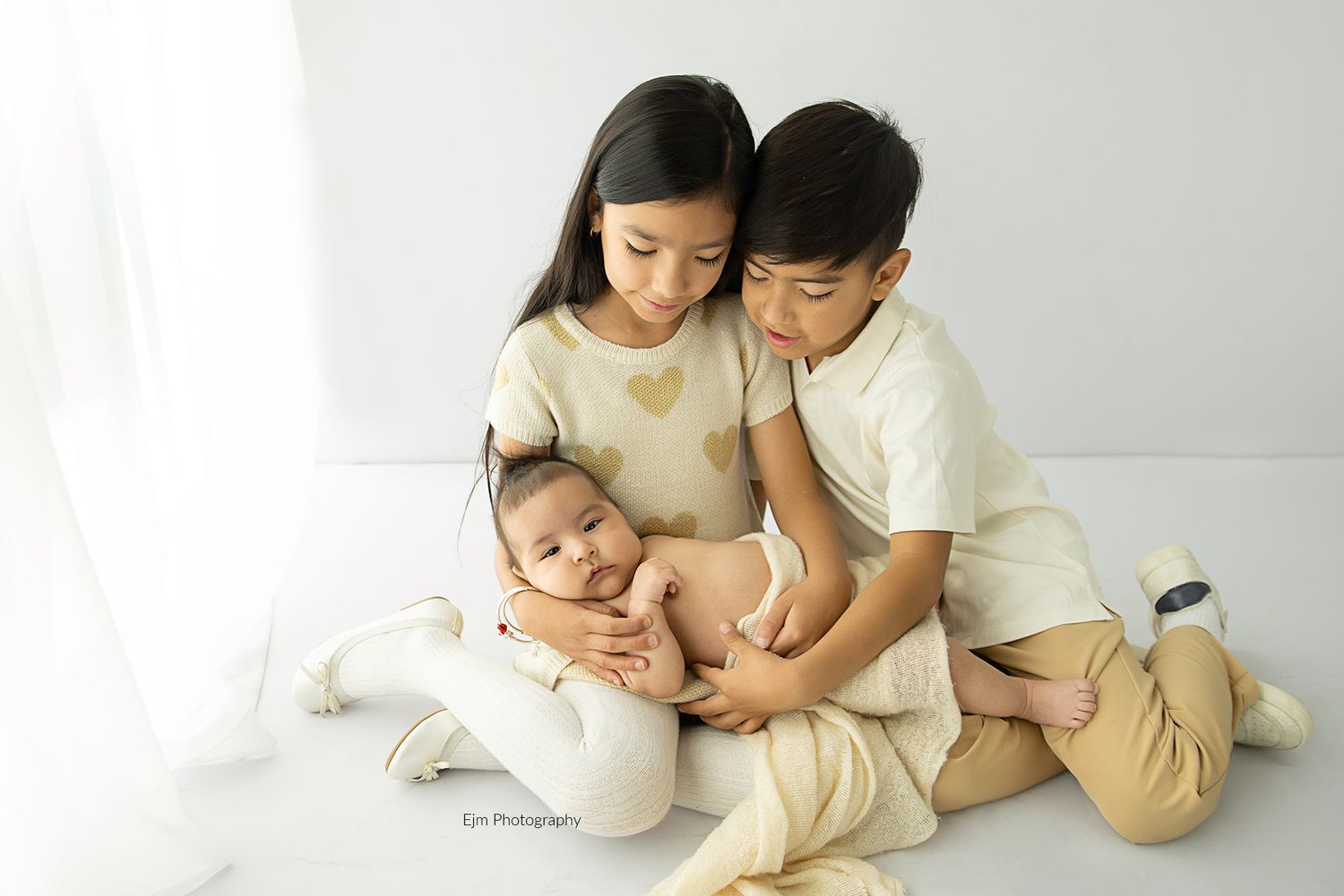 Sibling photo all in white and being. Two older siblings holding their new baby sibling in their laps.
