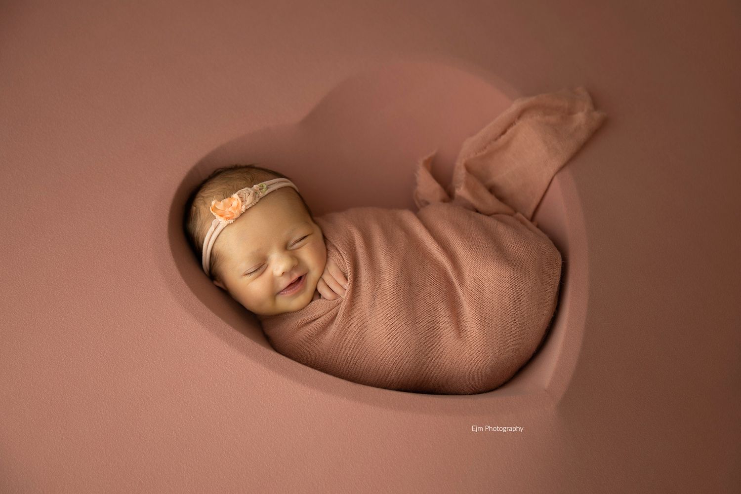 Baby girl in heart prop while wrapped giving a newborn smile.