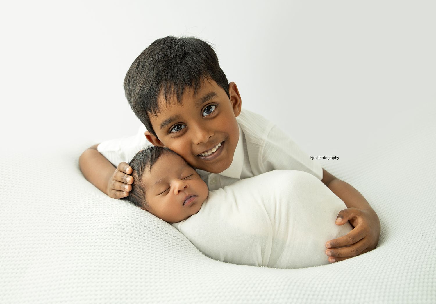 All while sibling photo. Newborn baby asleep and wrapped with big bother leaning on his head with a big smile, looking at camera.