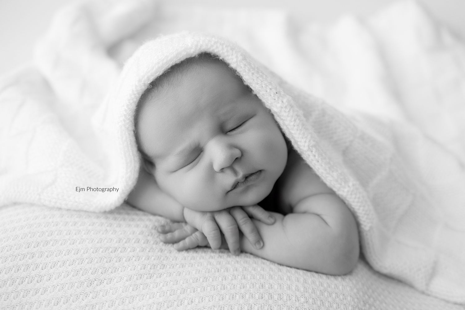 Black and white image of newborn baby sleeping with chin rested on hands and white blanker placed over his head with full face exposed.