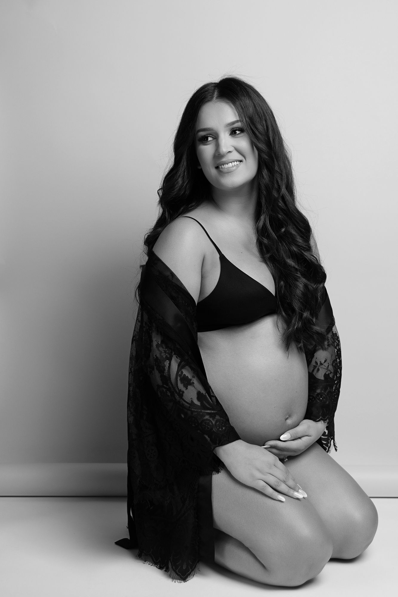 Pregnant lady sitting on her knees holding her baby bump, wearing a lace robe.