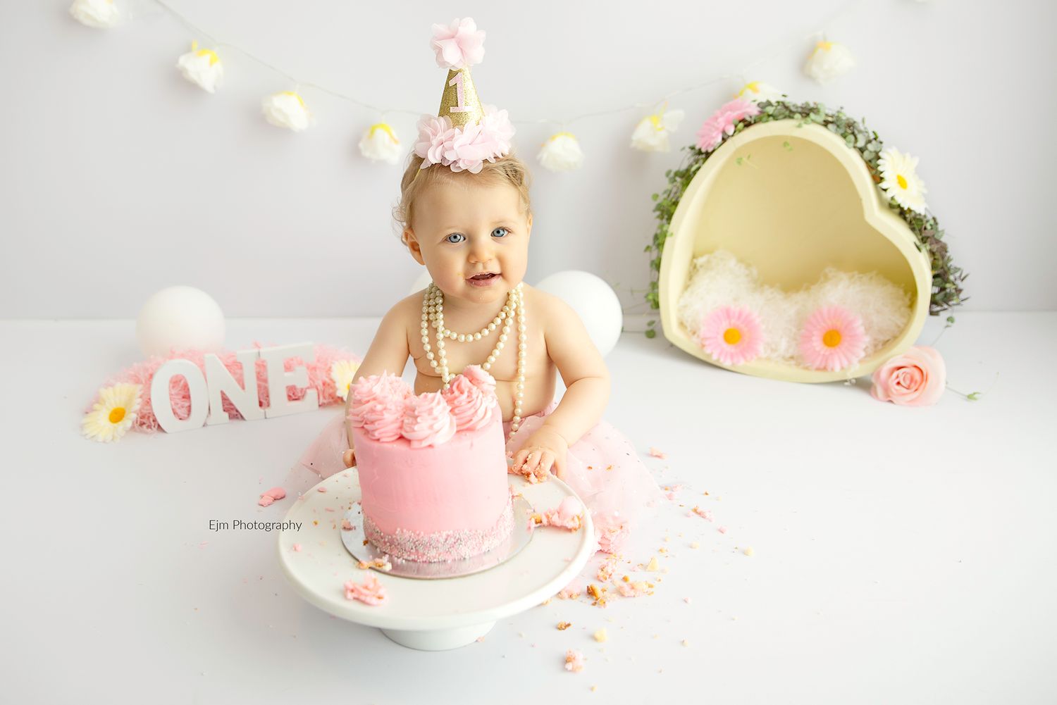 Baby girl in a girly setup of flowers, pearls, hearts and a pink cake for cake smash photo.
