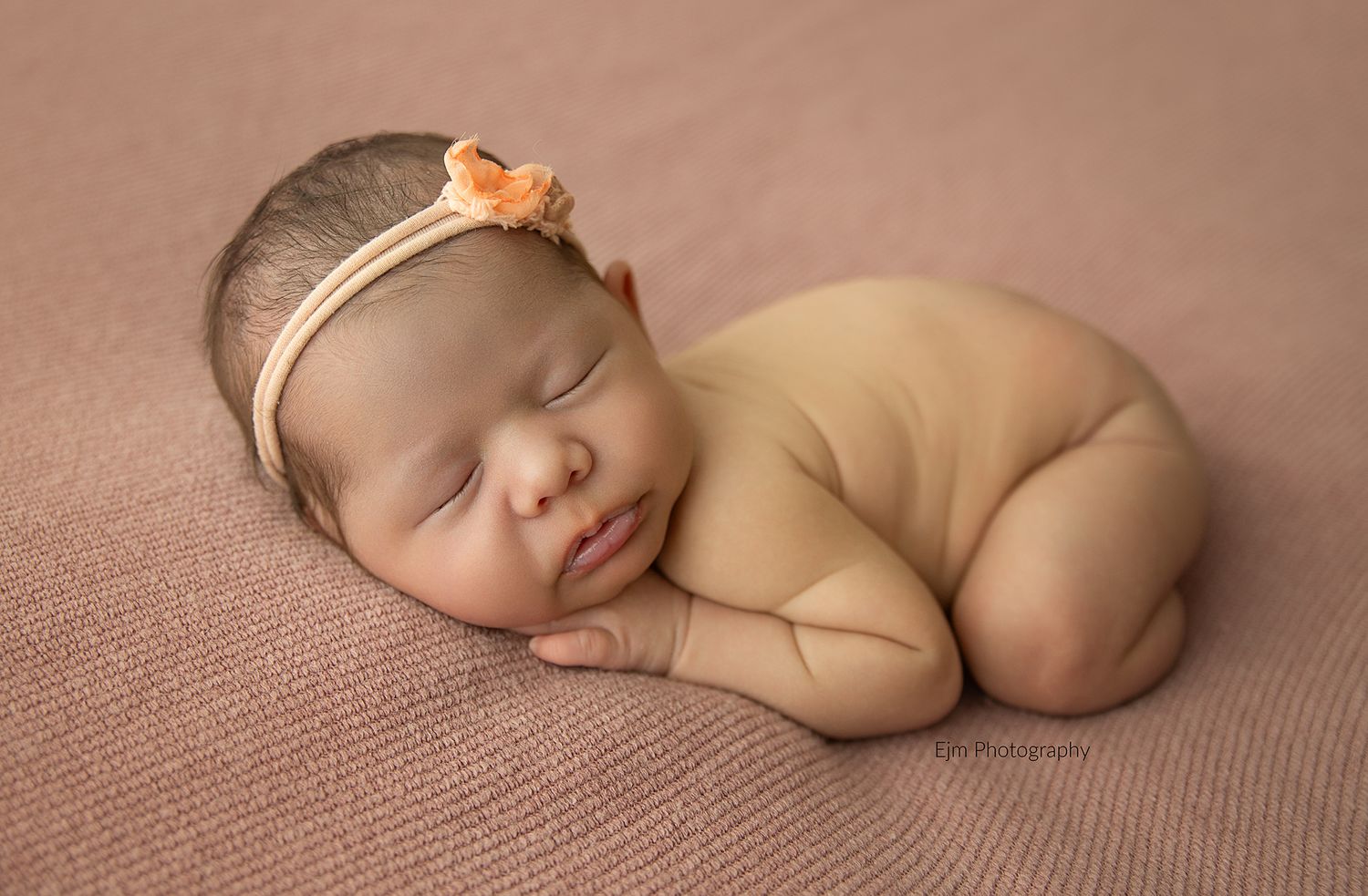 Newborn baby girl posed with her chin under hands, sleeping peacefully on pink blanket. Wearing a cute girly headband.
