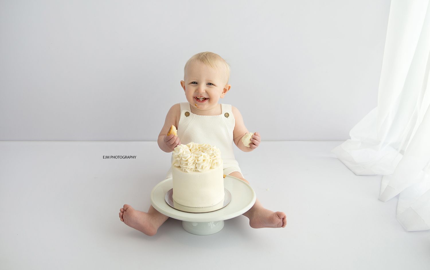 Baby boy in all white cake smash setup. Wearing cute white knitted overalls.