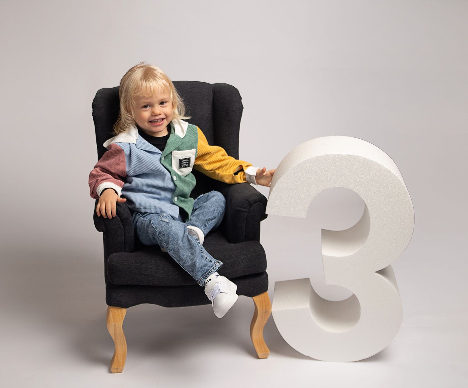3  year old blonde boy wearing colourful jacket sitting on little grey chair next to a 3 prop.