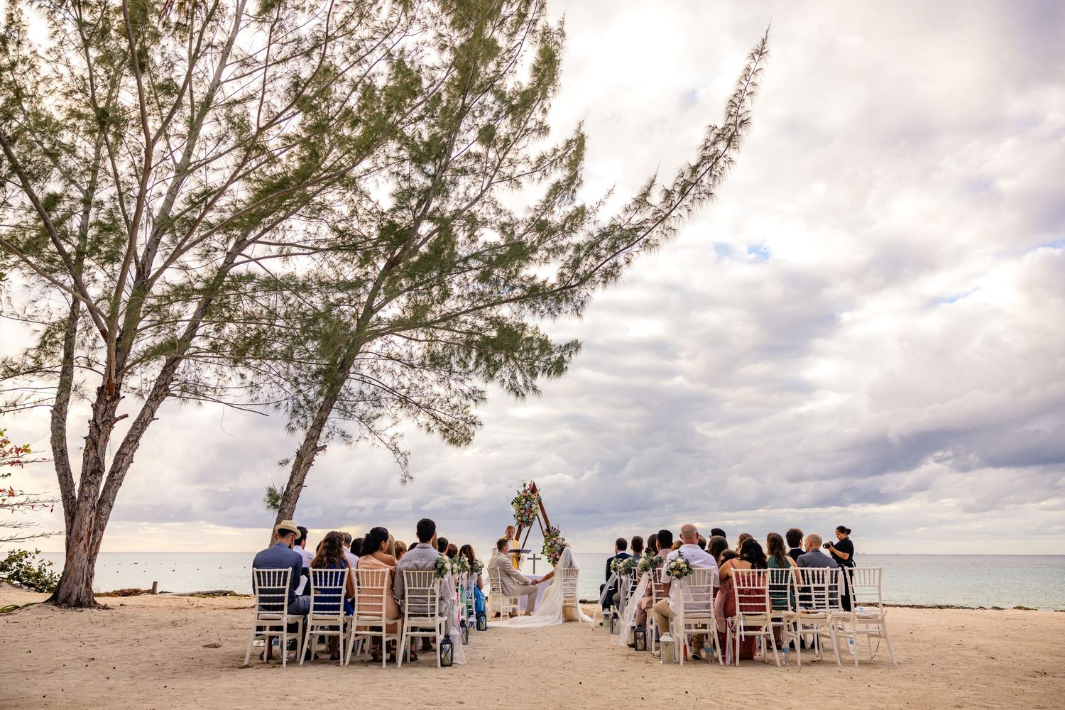 A beachside wedding ceremony setup with white chairs under a cloudy sky and palm trees by the ocean.