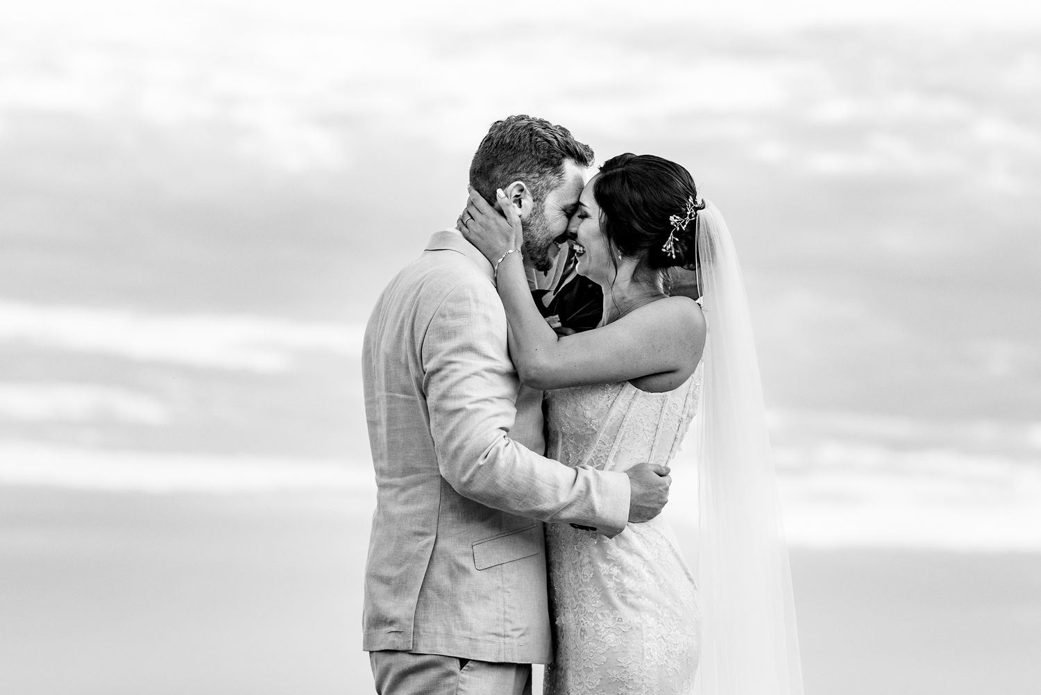 Black and white photo of a bride and groom embracing and smiling at each other on a beach on their wedding day.