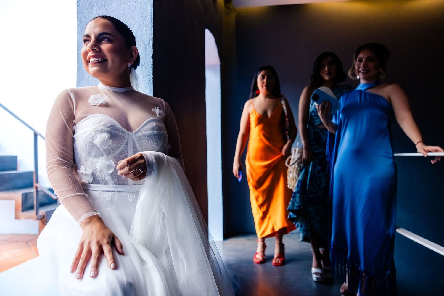 Bride in sheer white gown smiles as bridesmaids in orange and blue dresses walk behind her indoors.
