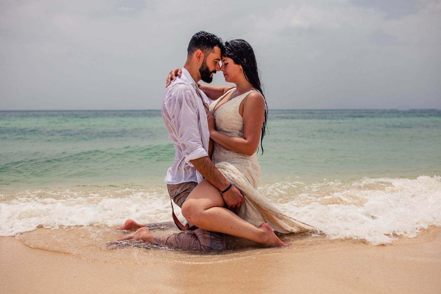 Romantic couple embracing on a tropical beach shore with turquoise ocean waves in the background.