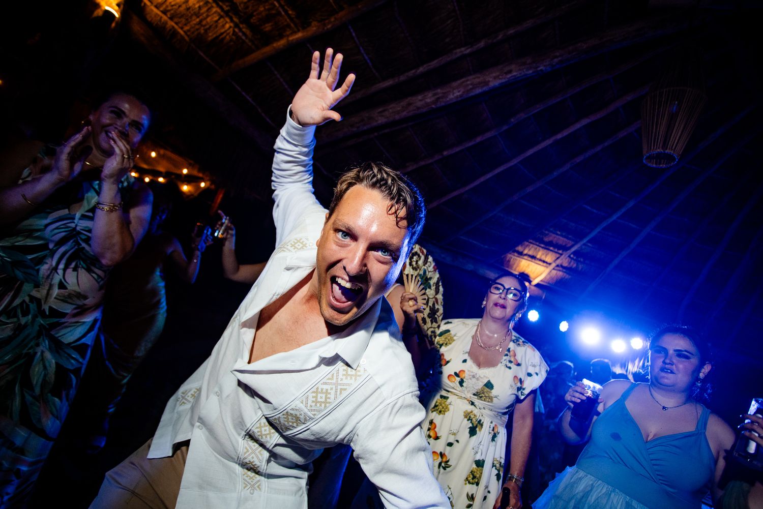 Energetic wedding guest dancing and raising hand at a lively indoor reception party with guests and string lights.