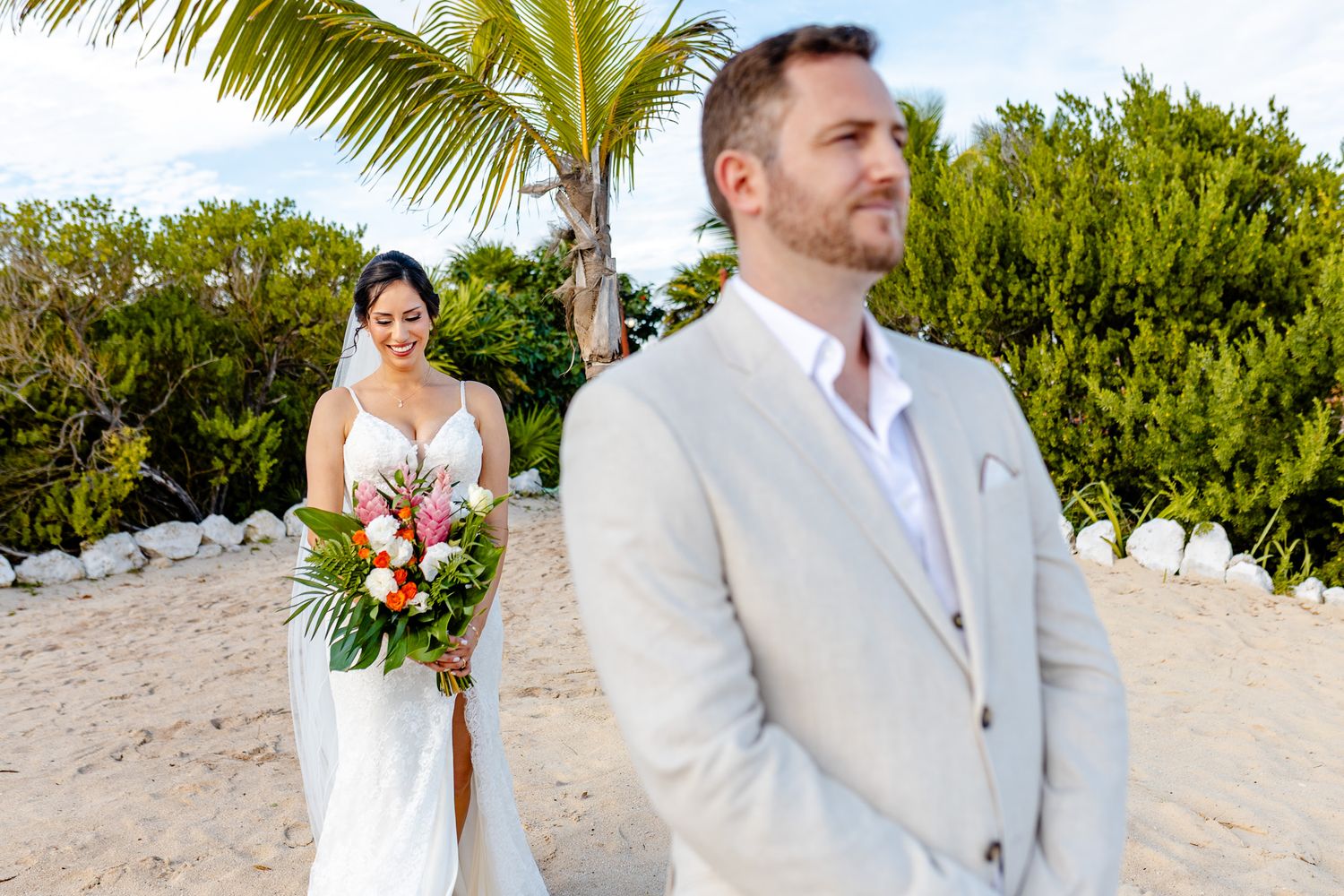 Bride in white gown holding tropical bouquet approaches groom in tan suit during beach wedding ceremony with palm trees.