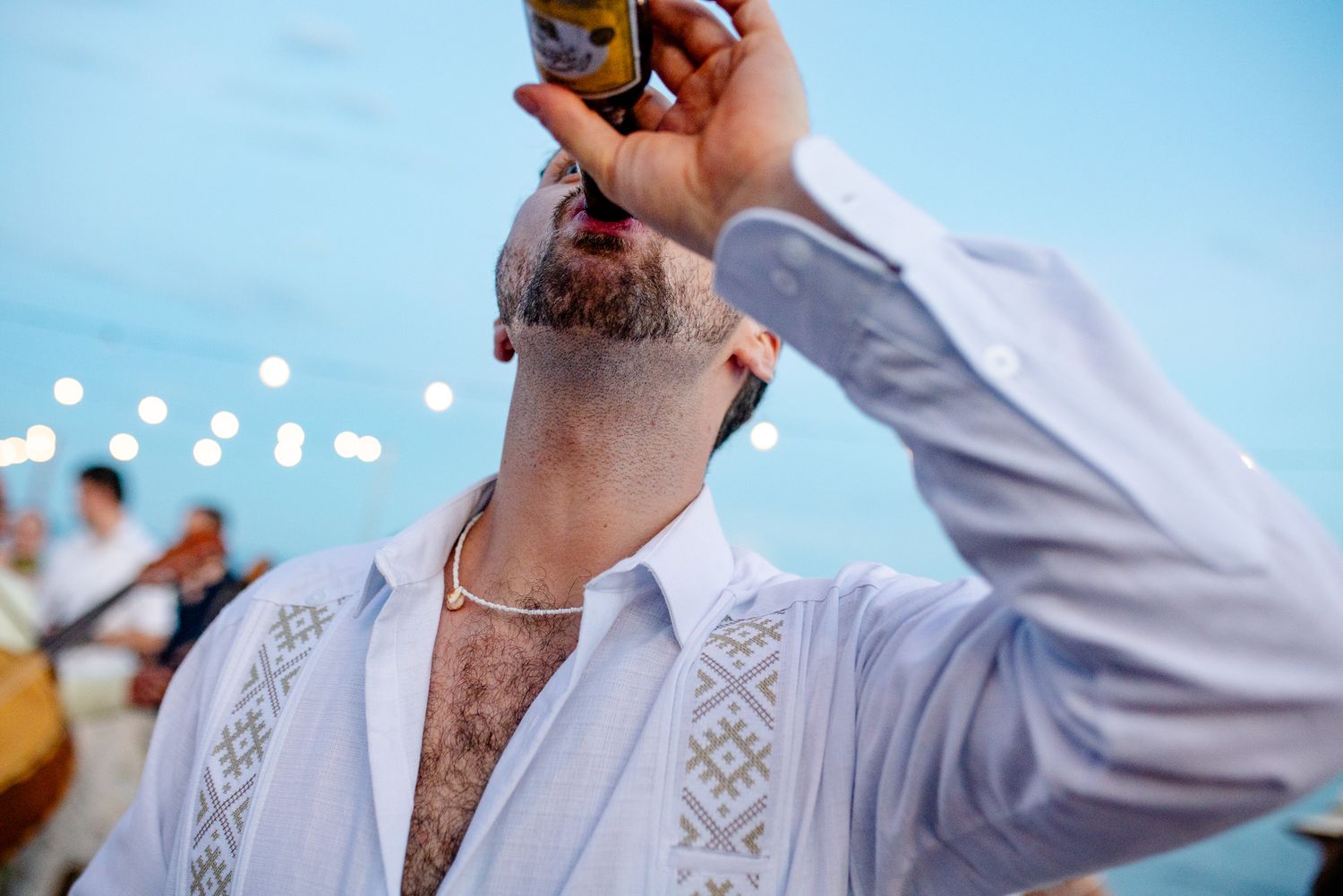 Man in white embroidered shirt drinking a Corona beer bottle at an outdoor beach celebration.