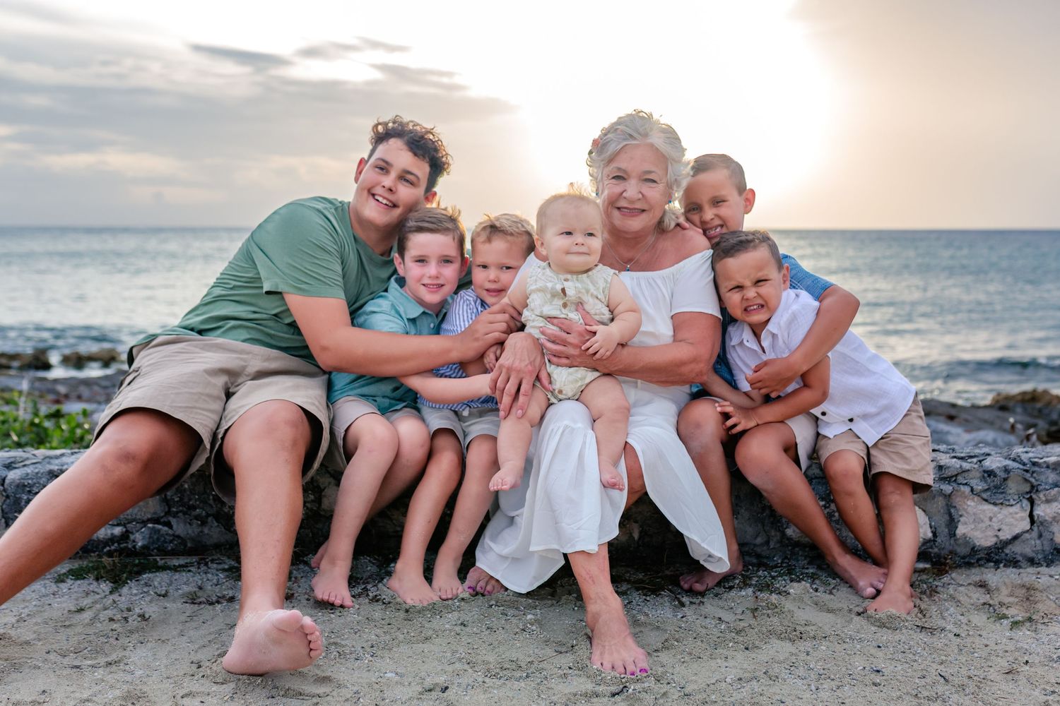 Family portrait of grandma with five grandchildren smiling together on a rocky beach at sunset.