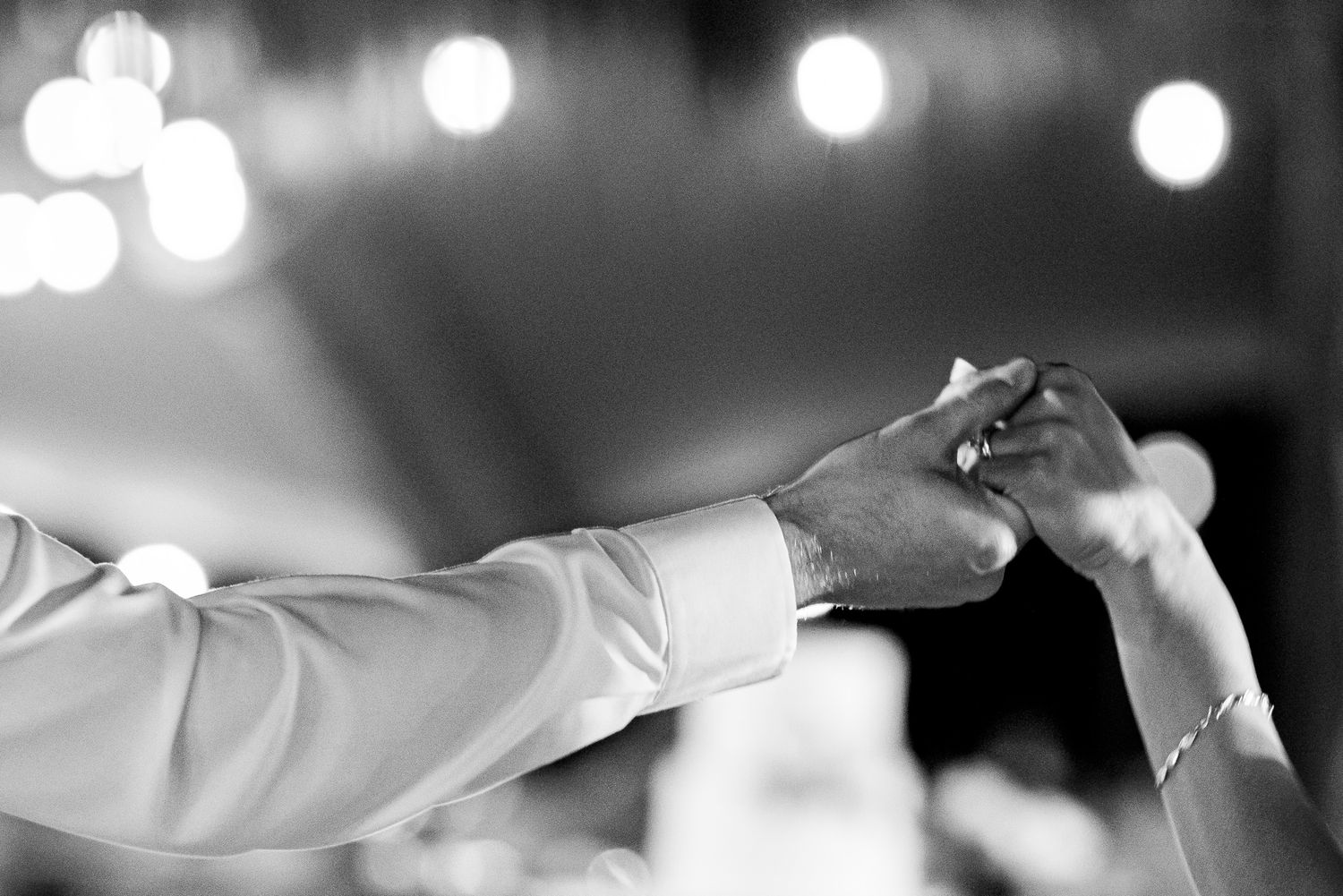 Black and white photo of a couple holding hands while dancing at a wedding reception with bokeh lights in the background.