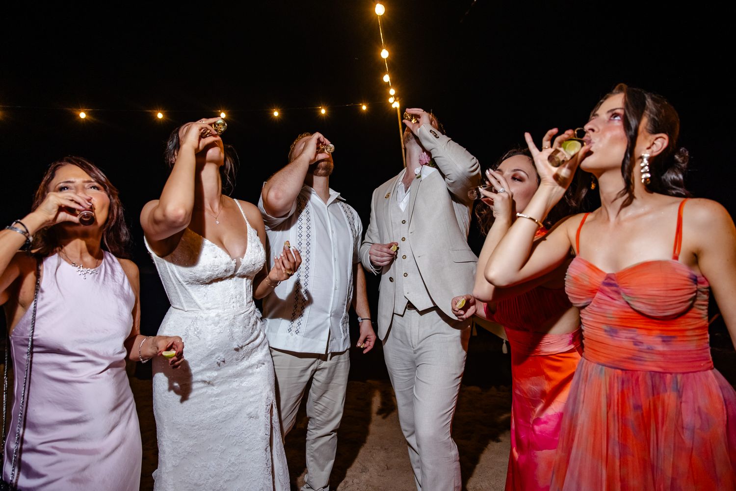 Wedding party guests doing shots with sparklers at nighttime beach celebration.