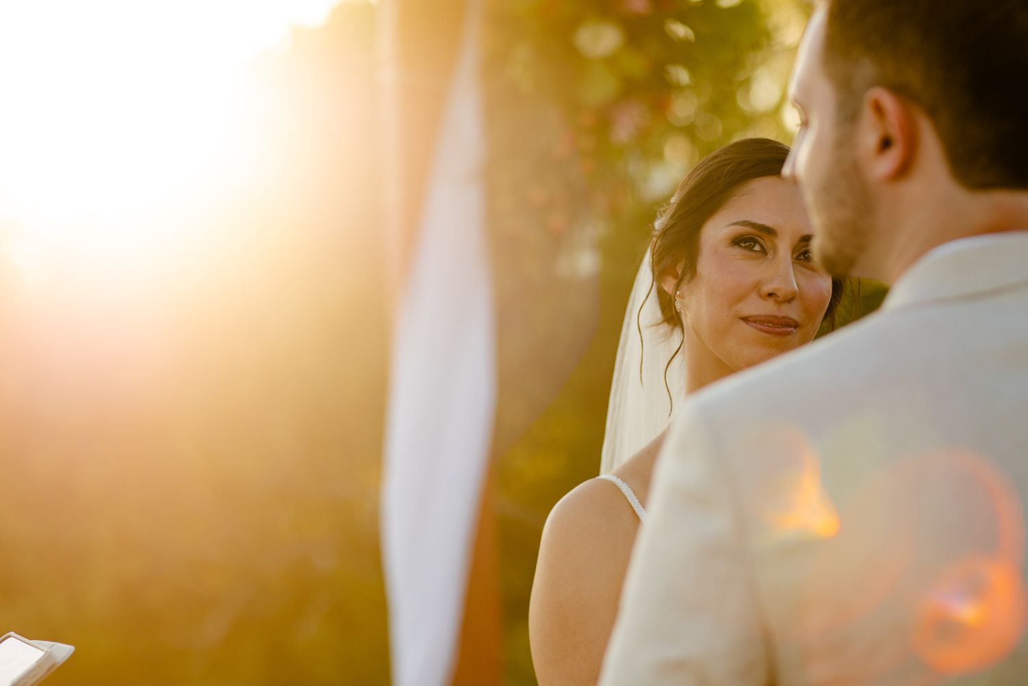 Bride gazing lovingly at groom during outdoor wedding ceremony with golden sunset light glowing in background.