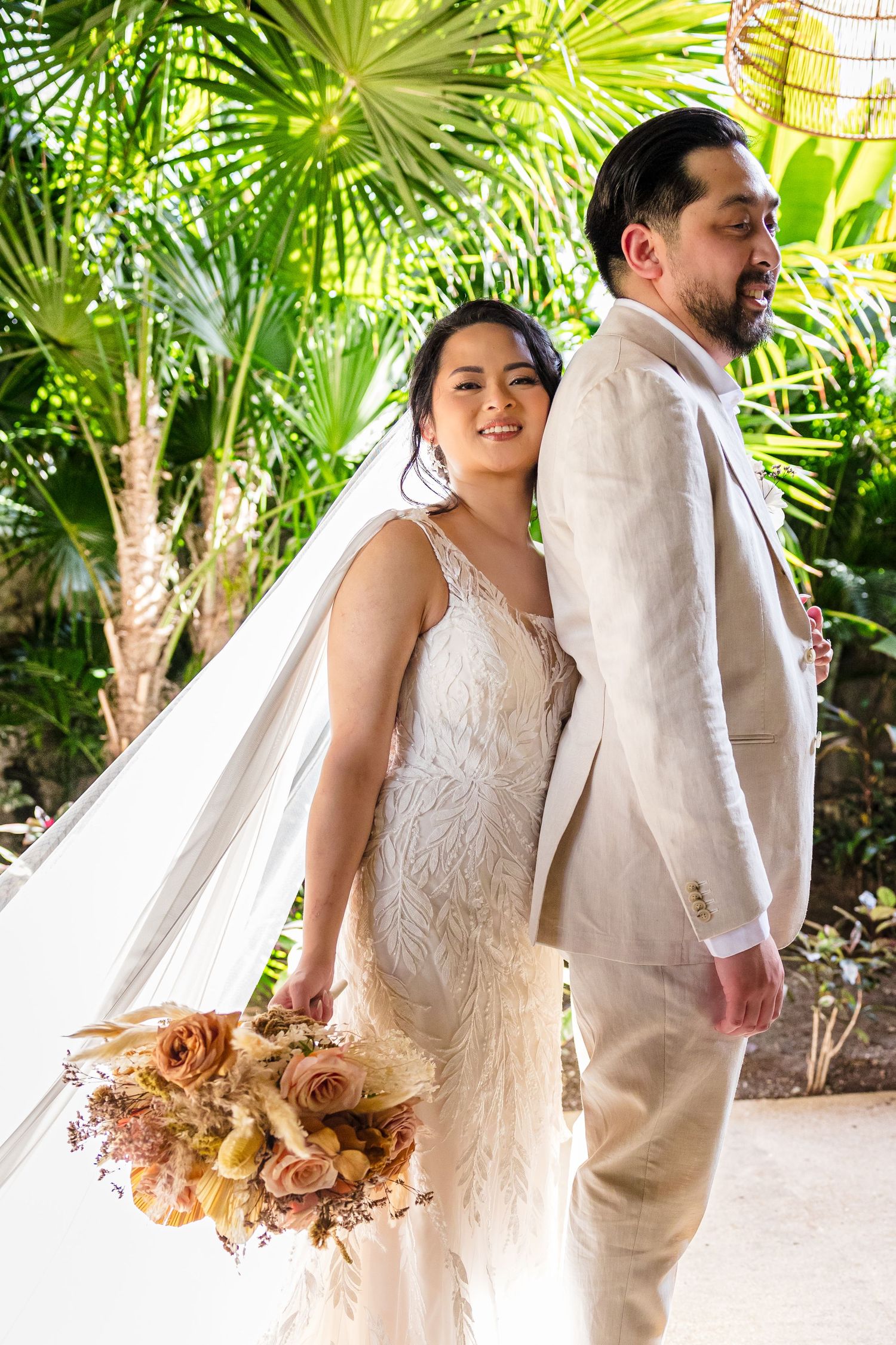 Bride in white feathered gown with bouquet stands back-to-back with groom in beige linen suit among tropical palms.