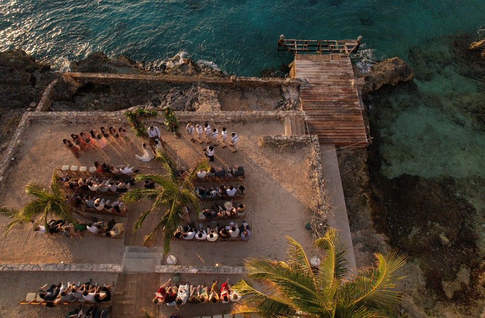 Aerial view of a beachside terrace with dining tables and palm trees overlooking turquoise ocean waters.