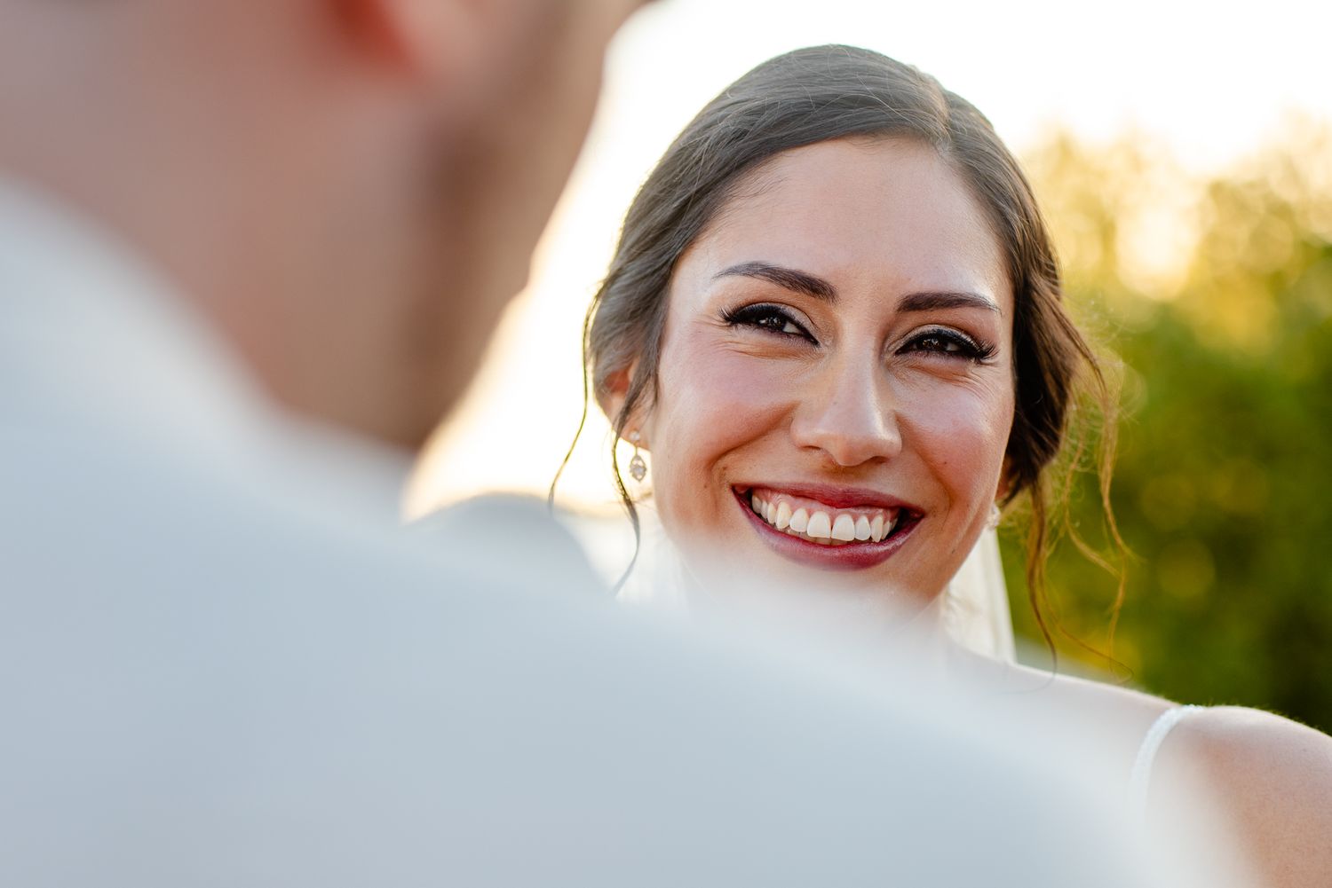 Smiling bride with elegant updo and dark lips gazes lovingly at groom during outdoor wedding ceremony.