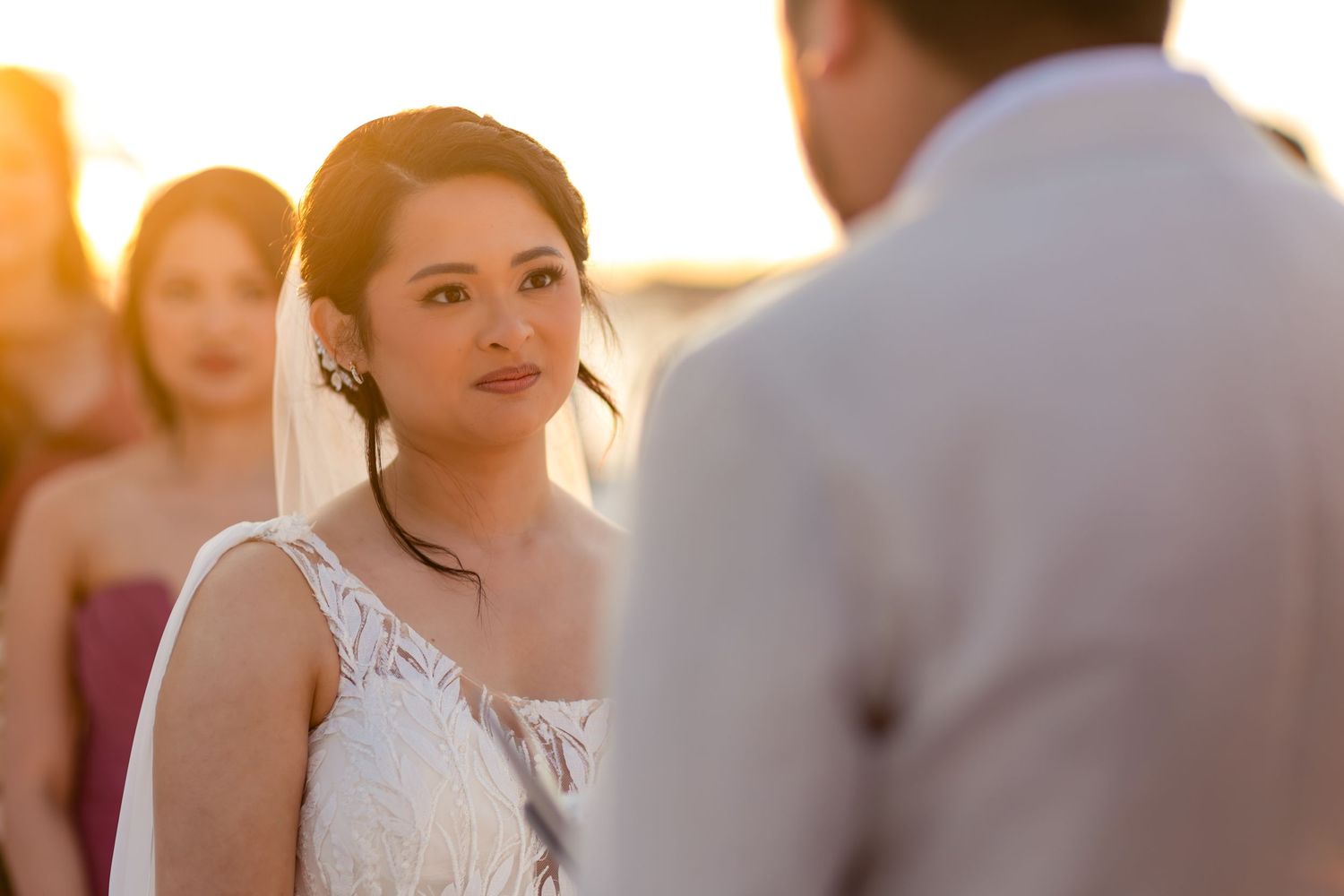 Bride in white lace veil gazes at groom during outdoor sunset wedding ceremony.
