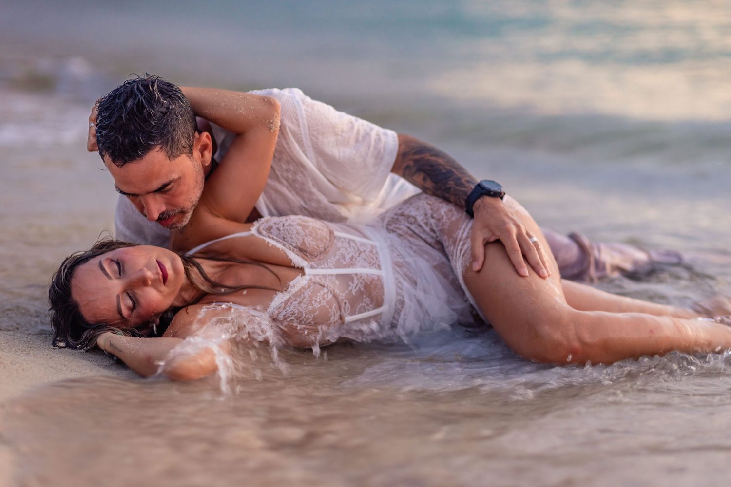 Romantic couple embracing in ocean waves on a beach, woman in white lace dress, intimate moment at sunset.