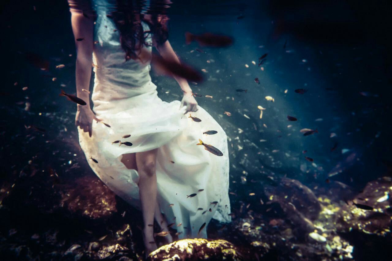 Woman in flowing white dress standing underwater surrounded by small fish in dark blue water.
