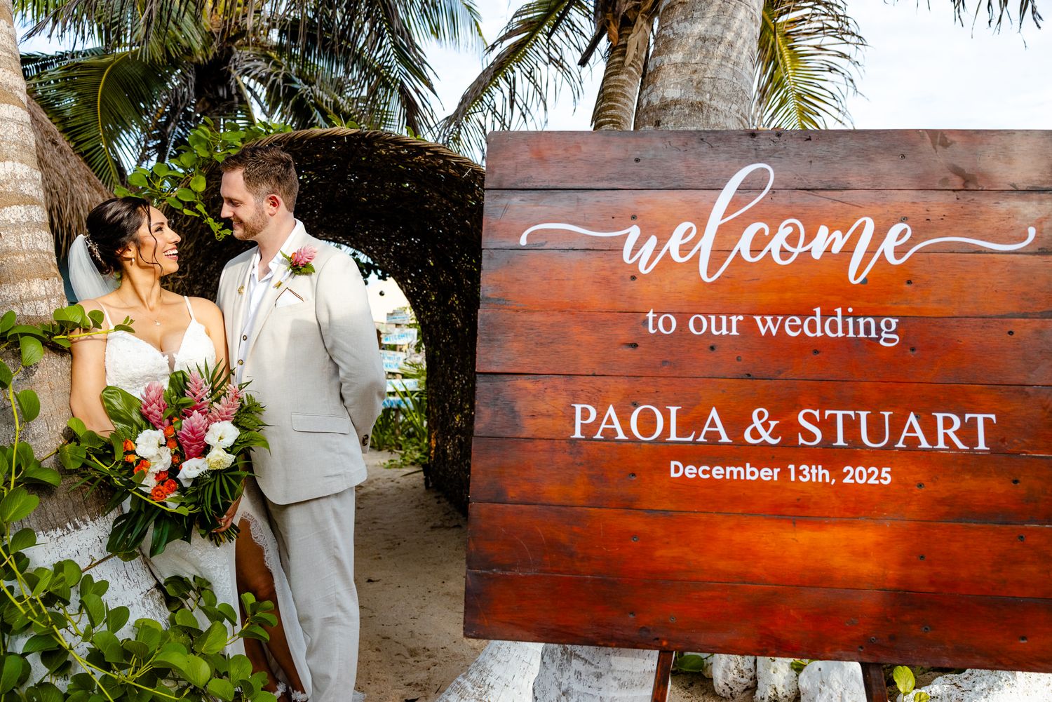 Bride and groom pose beside rustic wooden welcome sign for Paola and Stuart wedding on December 13th 2025.