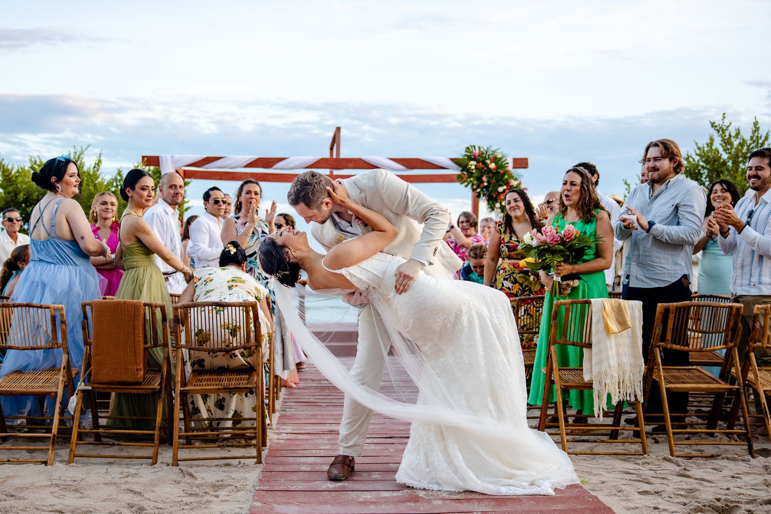 Groom dips bride for a romantic kiss at outdoor beach wedding ceremony with guests cheering.