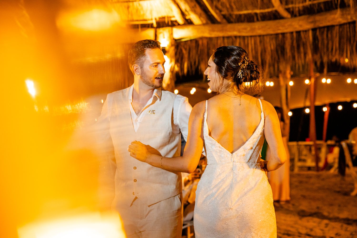 Couple sharing a romantic moment at a tropical beach wedding reception under warm string lights and a thatched roof.