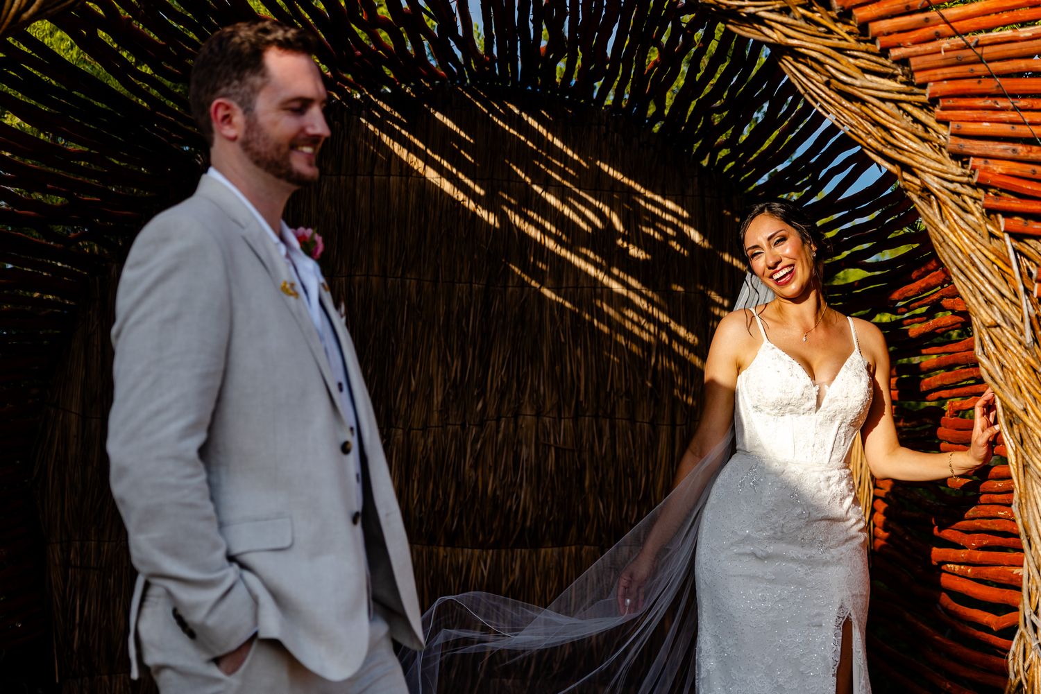 Bride in white gown and groom in tan suit smile together under a woven palm arch at a tropical wedding.