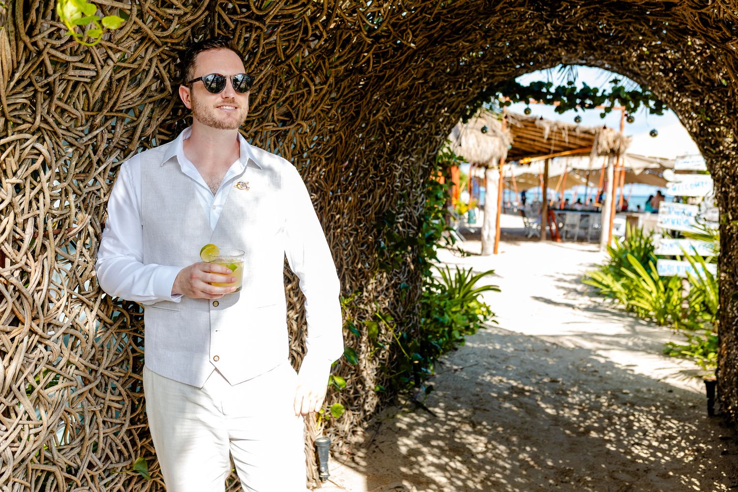 Man in white linen outfit holding a drink under a rustic twig archway at a tropical resort.