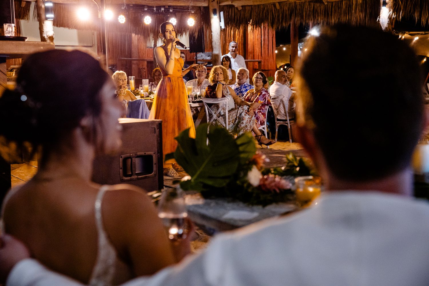 Woman in yellow dress giving a toast speech at an outdoor tropical wedding reception with string lights.