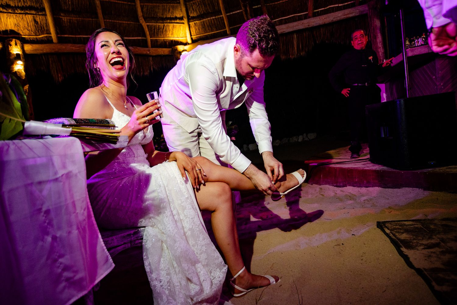 Bride laughing joyfully as groom puts on her shoe during a lively wedding reception celebration.