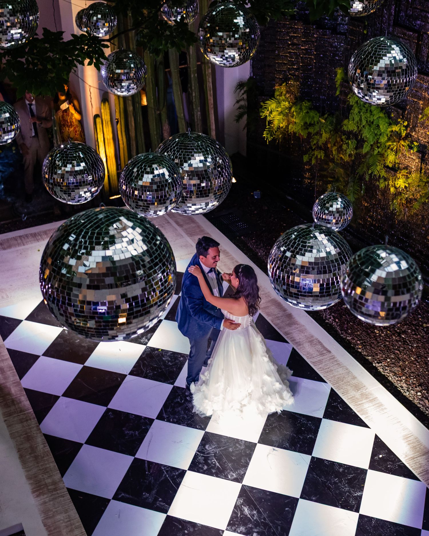 Bride and groom share first dance on checkered floor surrounded by hanging mirror disco balls at elegant wedding reception.