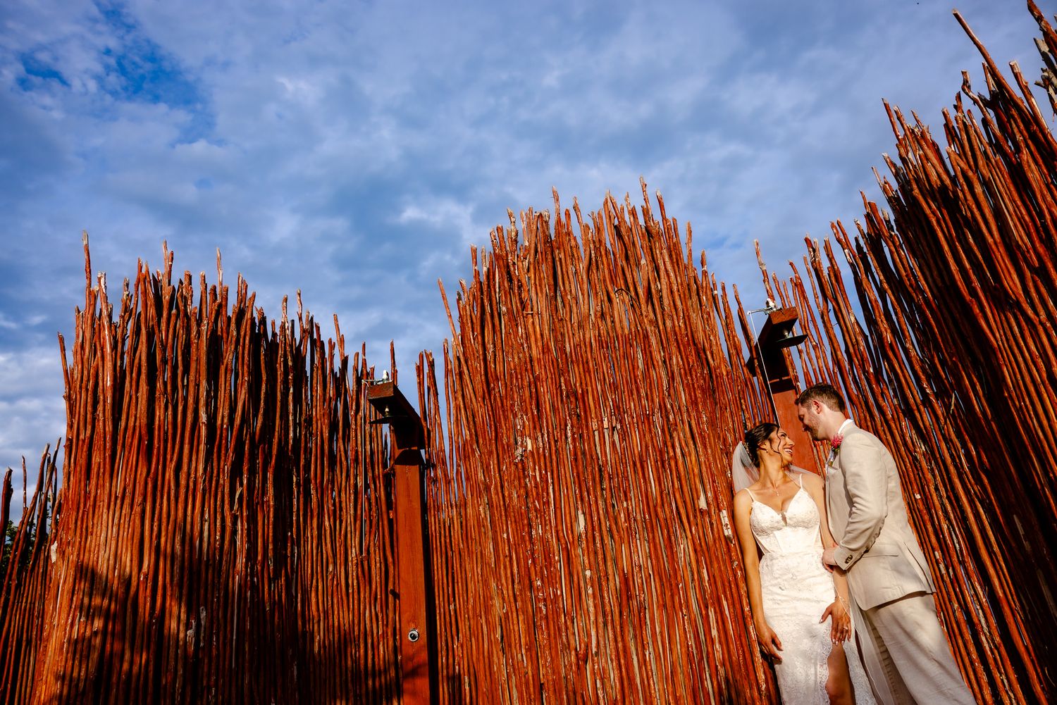 Bride and groom embrace near tall rustic reed fence under dramatic blue sky at outdoor wedding venue.
